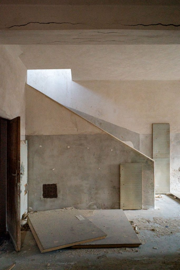 Bare room with a staircase rising on the right, pale peeling plaster, a panel leaning against the floor