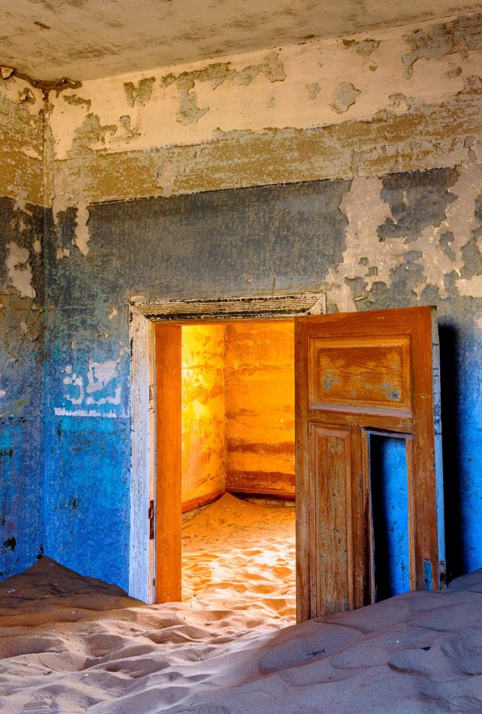 Room with pale blue walls and a door opening onto a space lit in orange, sand on the floor