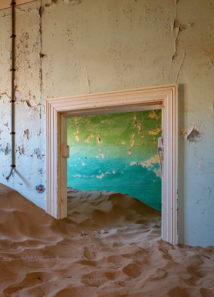 Doorway framing a peeling turquoise wall, with sand on the floor
