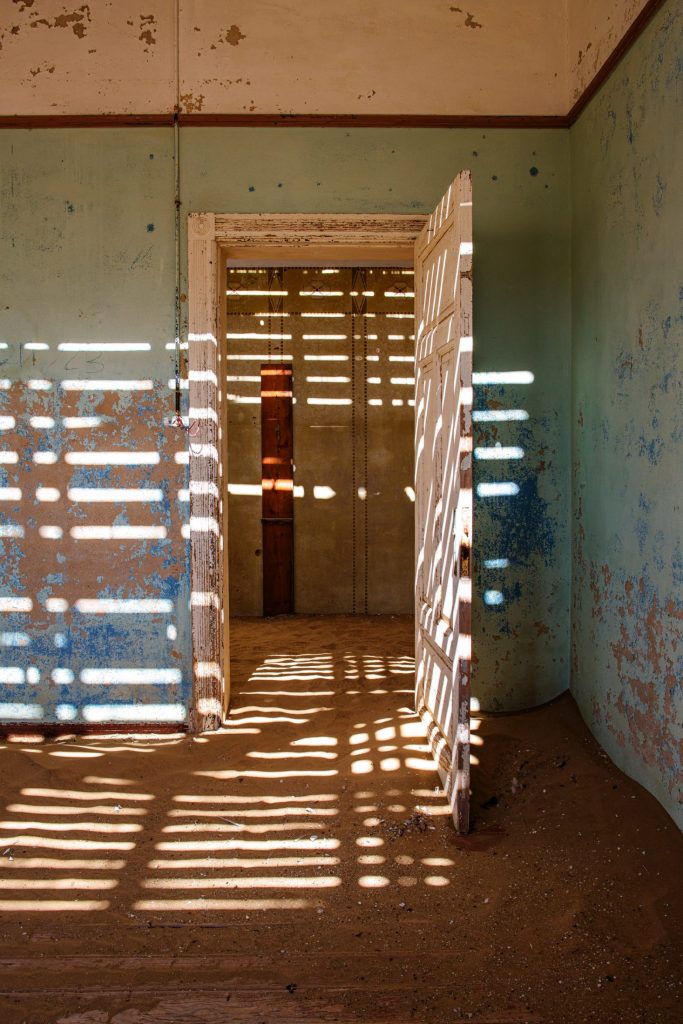 Abandoned room striped with light from slatted boards, with open doorways and sand on the floor