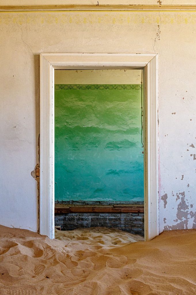 White door frame opening onto a wall washed in a green and turquoise gradient, sand on the floor